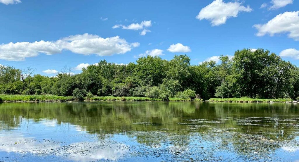 A calm river with green trees on the far bank, under a blue sky with scattered white clouds.