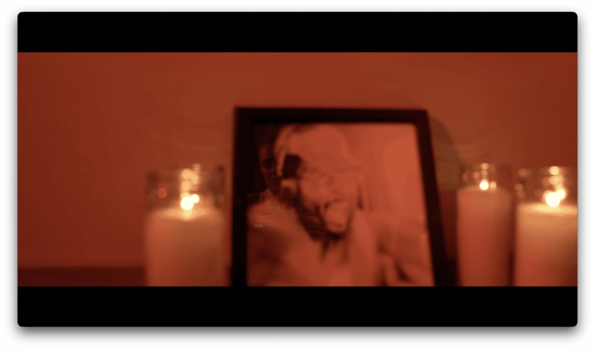 A blurry framed photo stands between two lit candles in a dimly lit, reddish room.