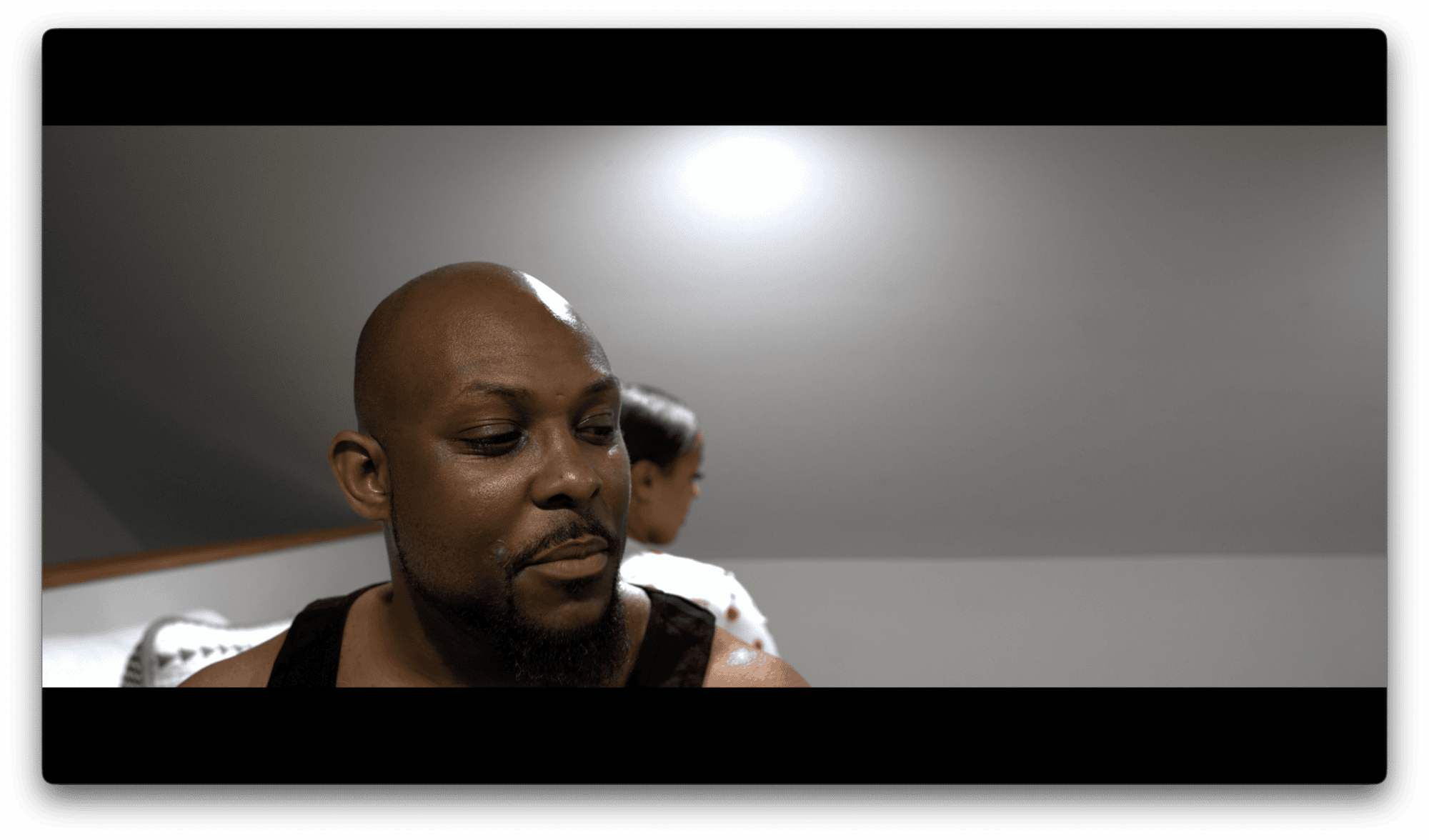 A man in a black tank top sits in the foreground looking down, while a woman in the background faces away from him in a dimly lit room.