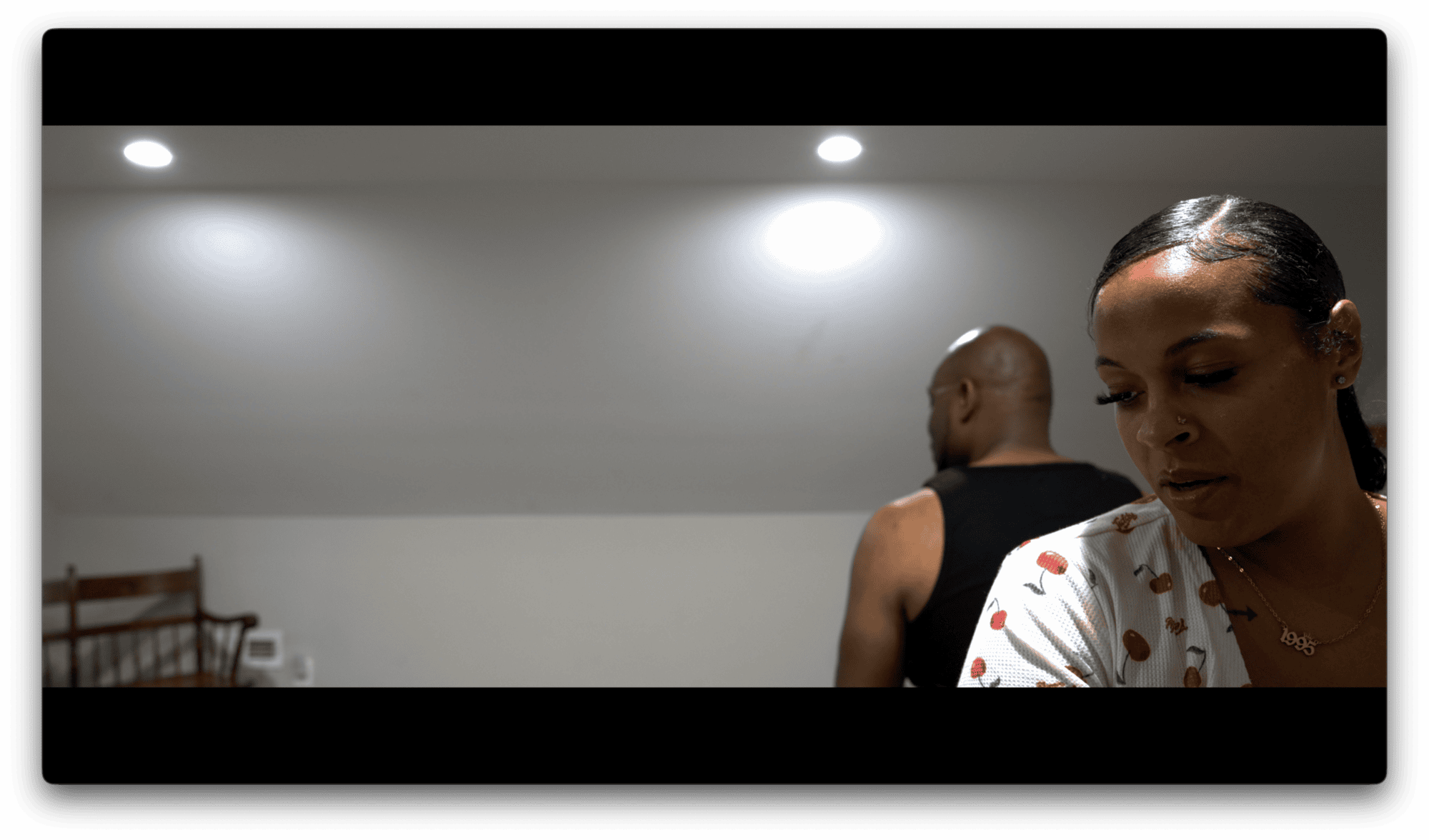 A woman in a white shirt stands in the foreground, looking down, while a man in a black tank top faces away in a dimly lit room.