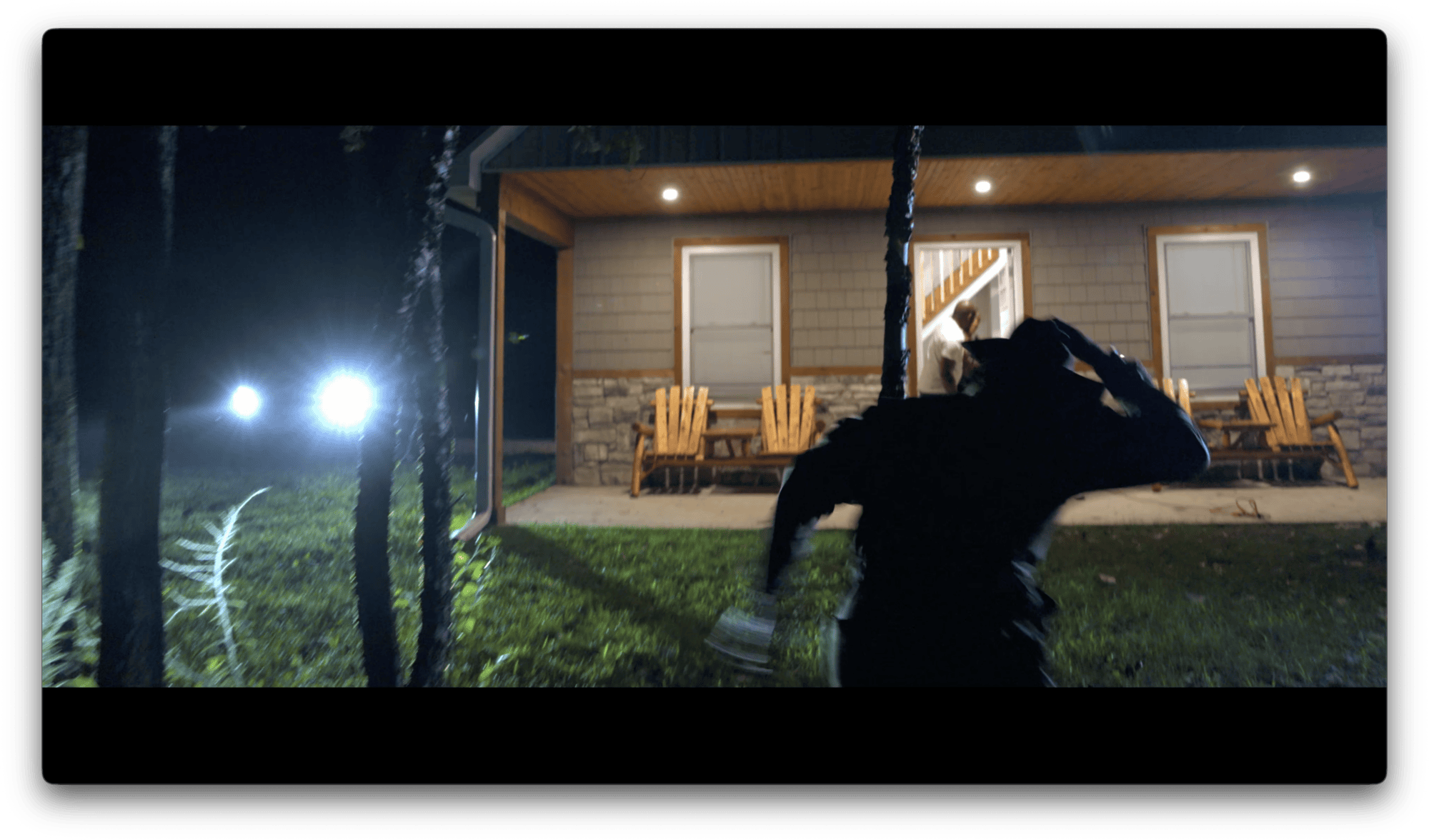 A person in shadow runs toward a porch with wooden chairs at night, illuminated by bright lights in the background.