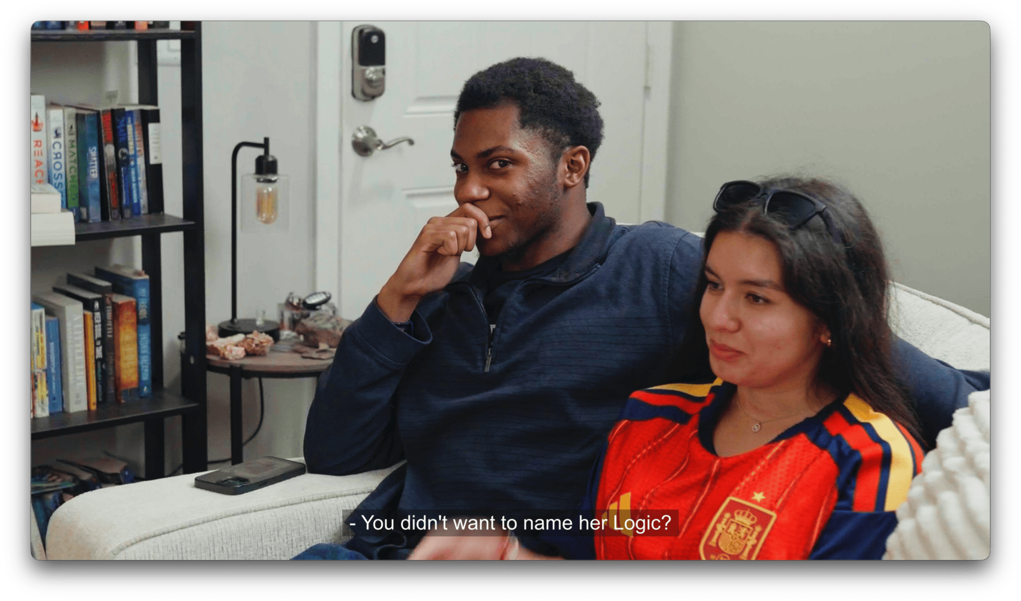 Two people sit together on a couch; the man is smiling with his hand near his mouth, and the woman is wearing a red sports jersey. Subtitles read,