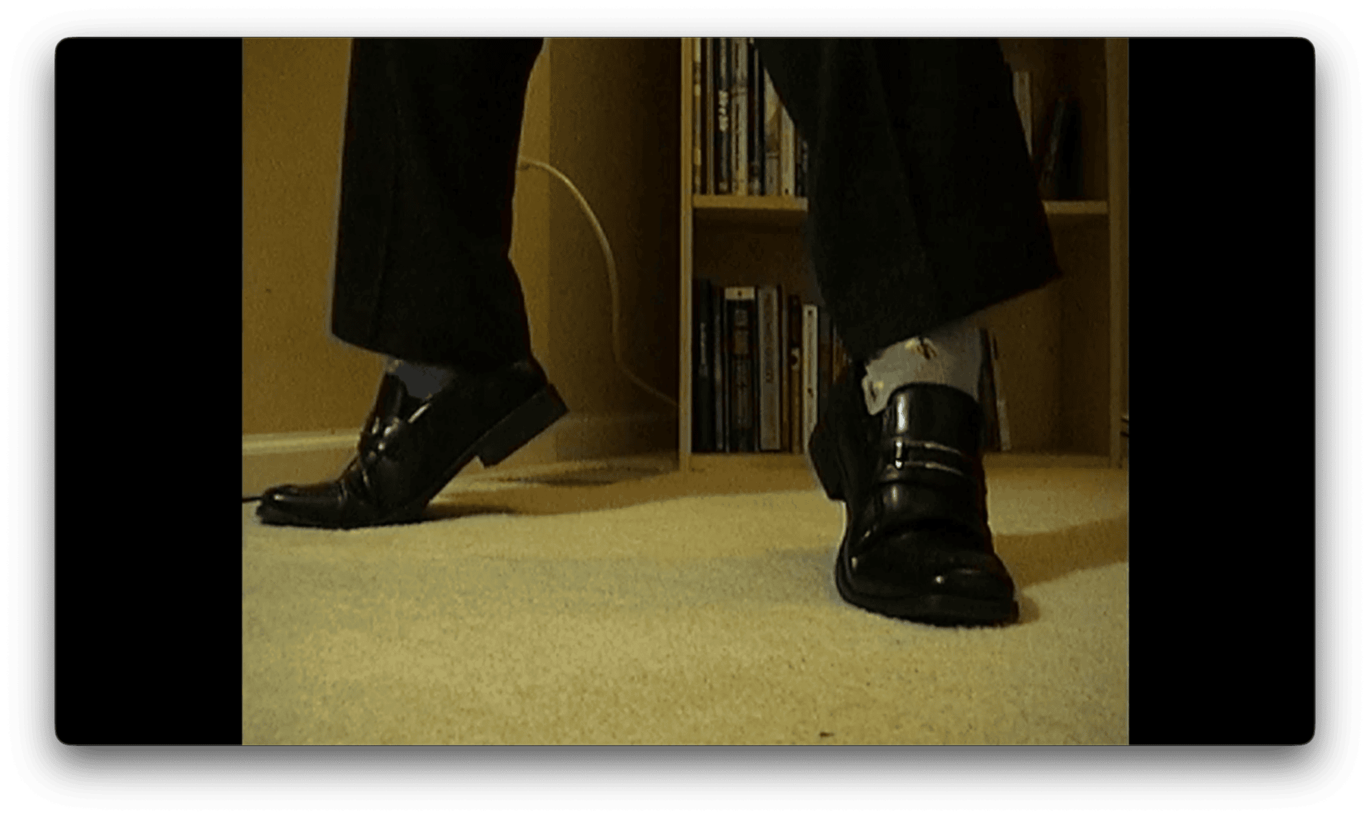 Two feet in shiny black dress shoes and mismatched socks stand on a carpeted floor, with a bookshelf in the background.