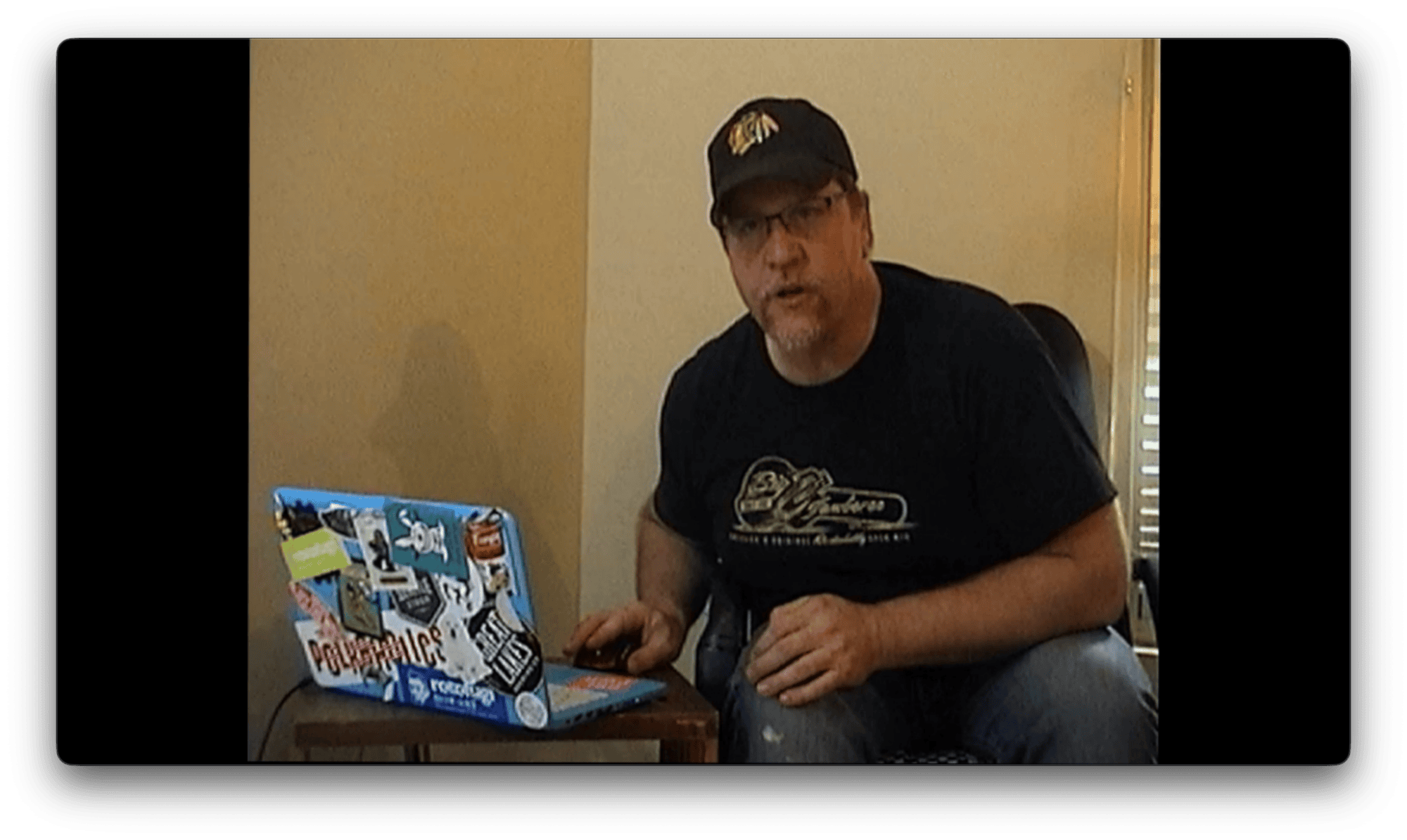 A man wearing a black cap and t-shirt sits next to a sticker-covered laptop, speaking to someone out of the camera's view.