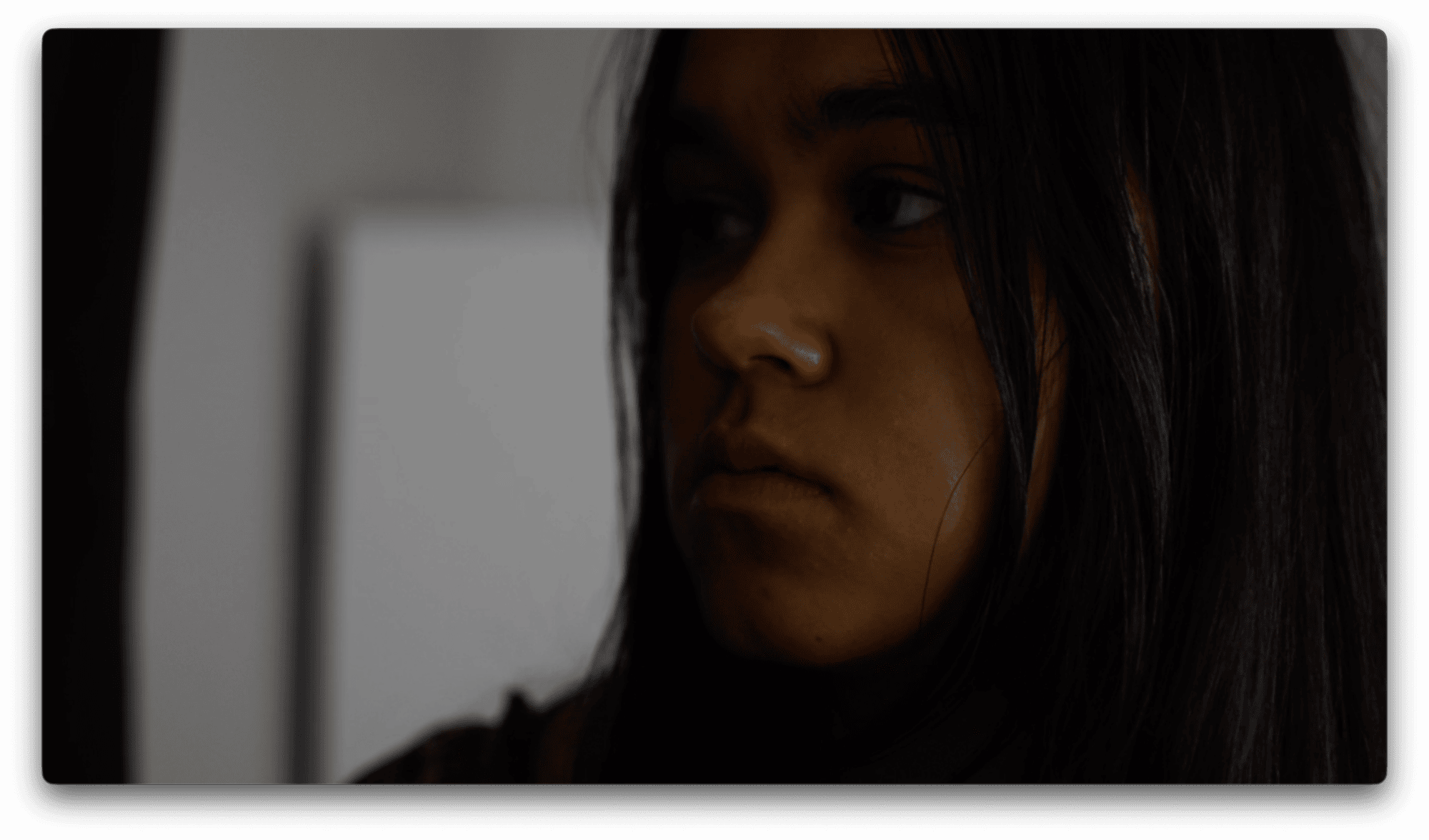 A person with long dark hair looks to the side in a dimly lit room, their expression neutral.