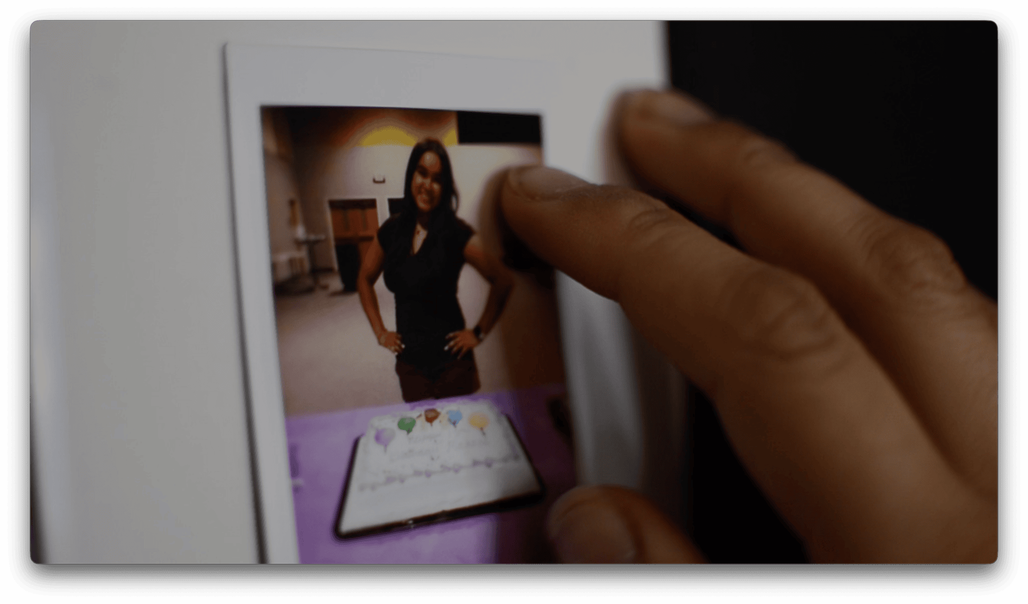 A hand points to a printed photograph of a woman standing indoors, with a decorated cake on a table in front of her.