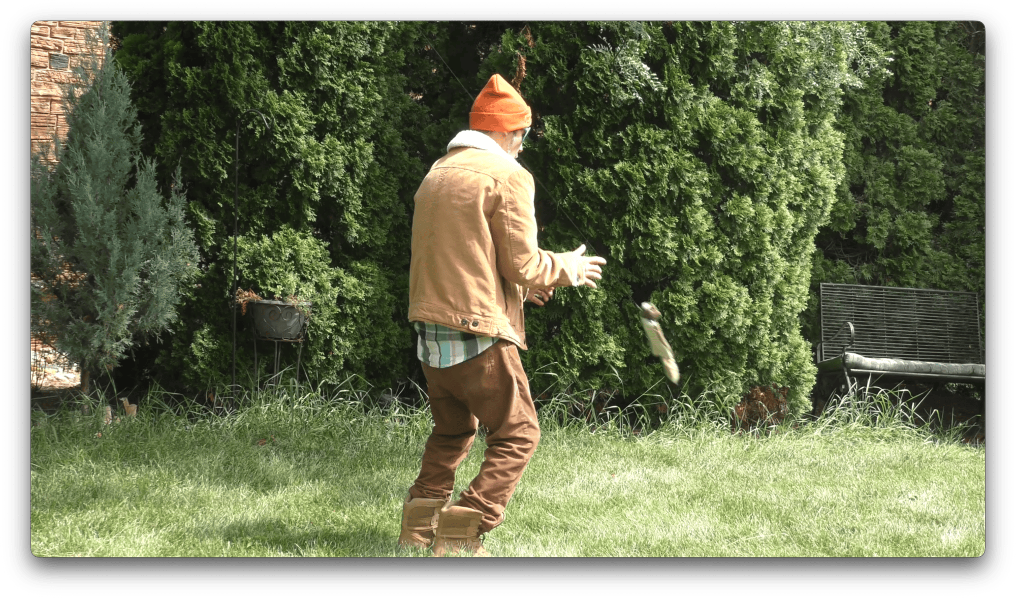 A person in brown clothes and an orange hat stands on grass, reaching. A stuffed squirrel is flung at them, carried across the scene by visible fishing line