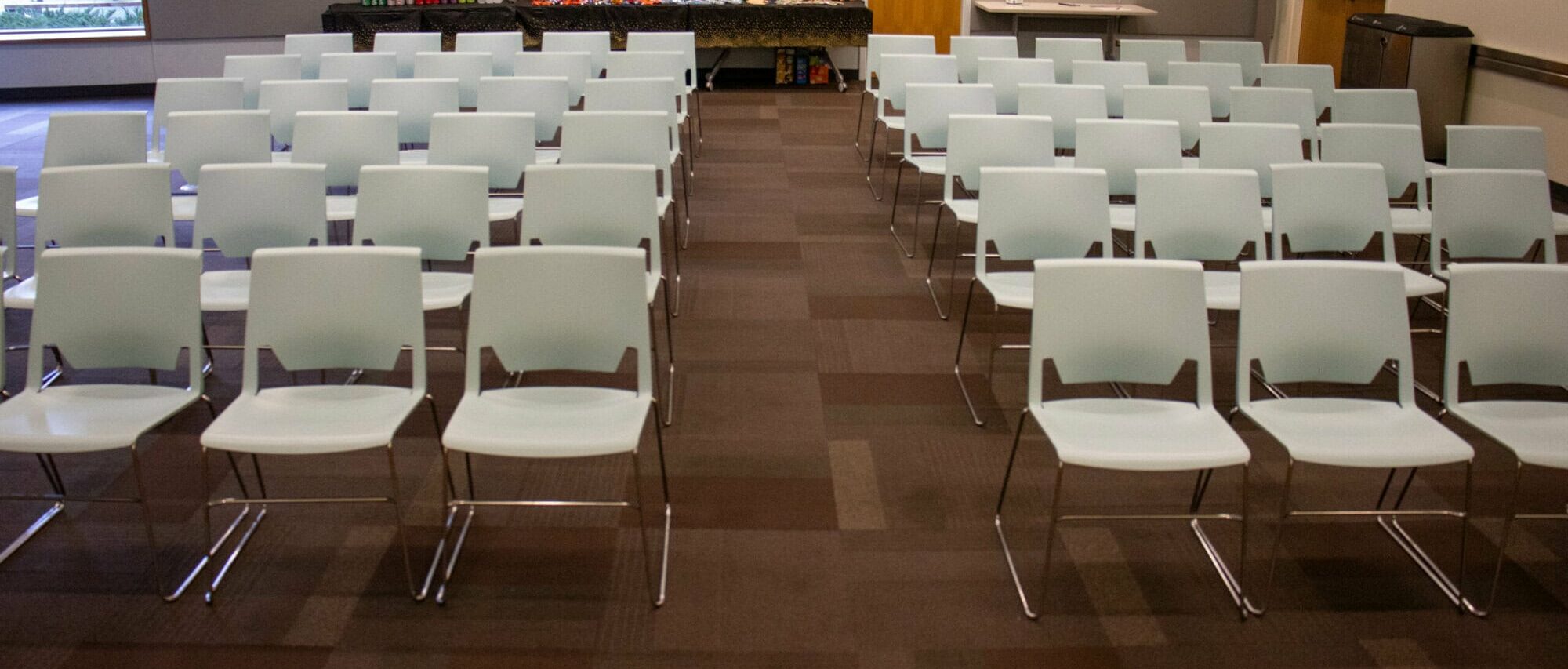 Rows of empty blue chairs in a conference room with a table of snacks and drinks at the back.