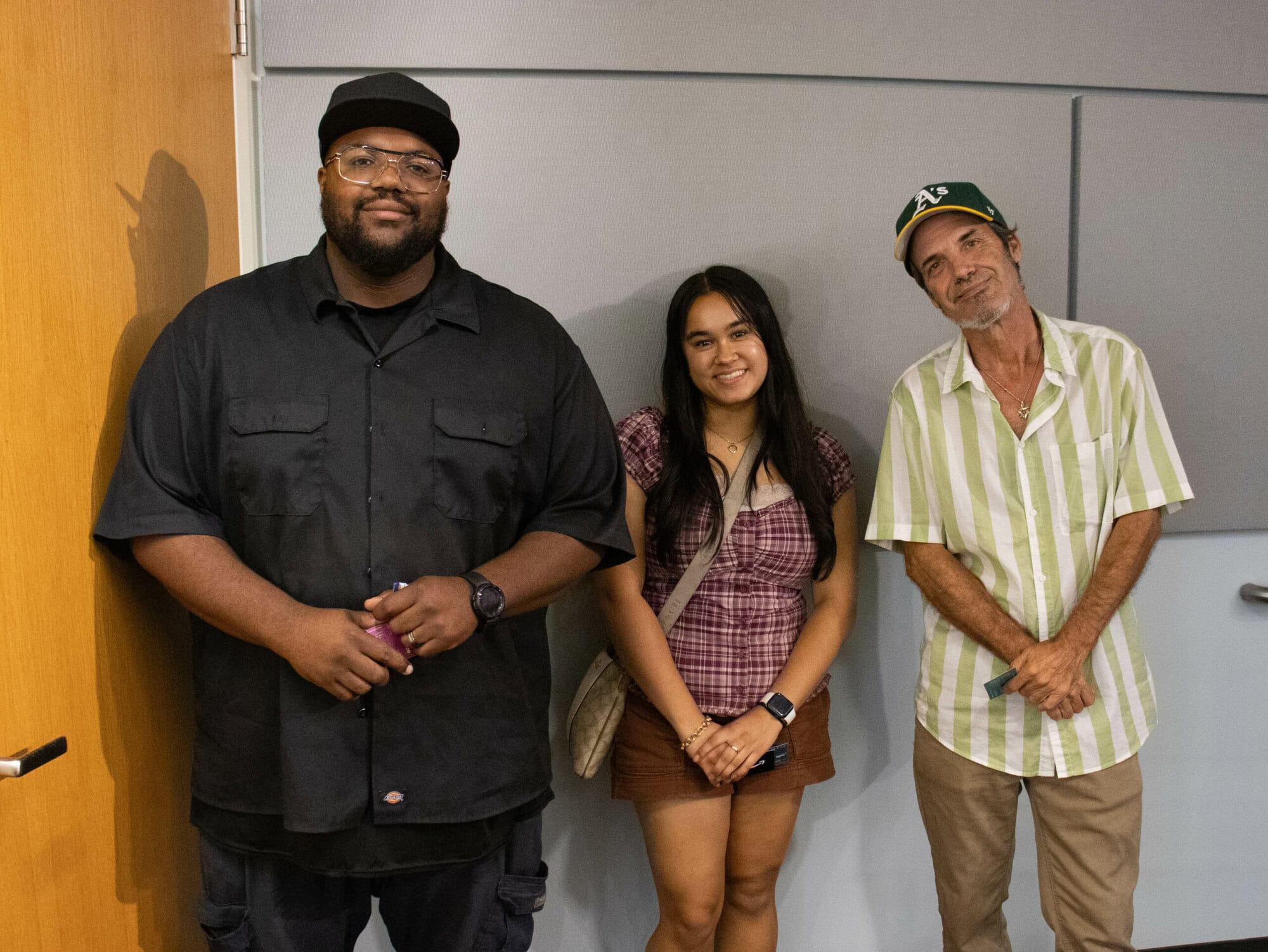 Three people stand indoors against a gray wall, each holding a ticket. One wears a black shirt and cap, another a plaid shirt and skirt, and the third a striped button-down shirt with a green cap.